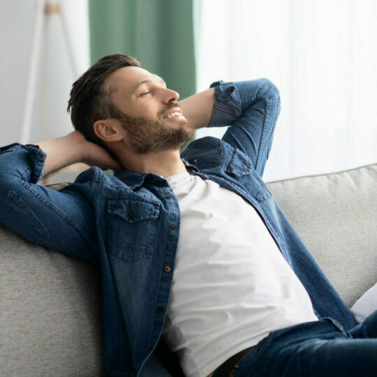 Ein glücklicher Mann sitzt mit zurückgelehnten Armen auf einem Sofa und lächelt